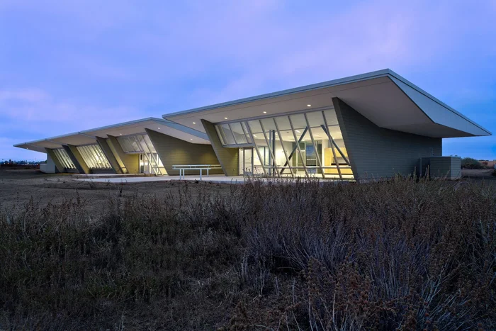 San Diego National Wildlife Refuge facility at dusk: winged roof planes over lit glazing, standing in low desert scrub.